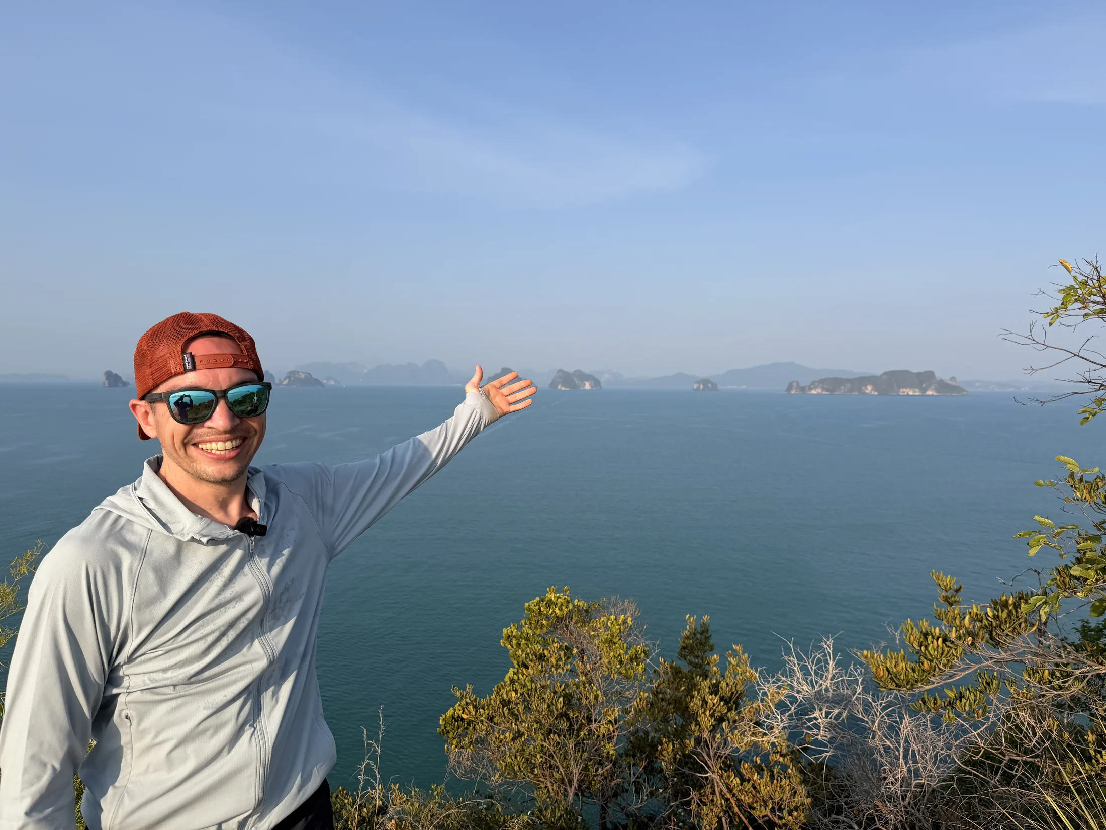Joe in an orange cap and sunglasses on a hilltop, arm out toward limestone islands in a blue sea in southern Thailand