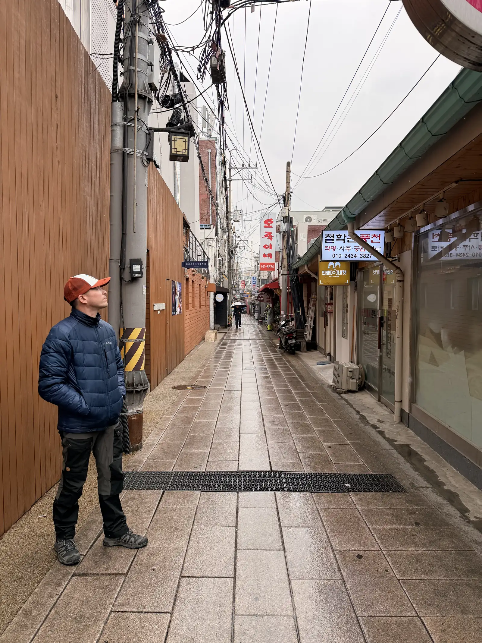 Joe in an orange cap and navy puffer jacket on a rain-wet street lined with shops in South Korea